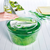 A person uses the Zyliss Swift Dry Salad Spinner Large, featuring a green and white design, clear bowl, and non-slip base on a white table. Green utensils, a yellow-striped napkin, bottles, and salad dishes are visible in the background.