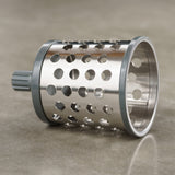 A stainless steel cylindrical grater drum with large round holes and a gray plastic attachment, resting on a smooth, light-colored surface.