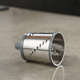 A cylindrical stainless steel rotary grater blade with sharp diagonal slots lies on a smooth, beige countertop. The blade has gray plastic edges and is designed for use in a kitchen appliance.