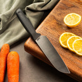 #15cm/6"
A Zyliss Santoku Knife with an ergonomic handle sits on a wooden cutting board beside three lemon slices, a halved lemon, two whole carrots, and a green cloth spread on the gray surface.