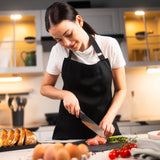 #15cm/6"
A woman in a black apron slices raw chicken breast with a Zyliss Santoku Knife on a cutting board in a modern kitchen, surrounded by eggs, bread, tomatoes, and herbs.
