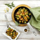 Brussels sprouts with bacon and herbs are perfectly cooked in the Zyliss Ultimate Pro Non-Stick Frying Pan With Pouring Lip, shown on a wooden board beside a served dish, black pepper, pine nuts, and rosemary atop a white wooden table. #20cm/8"