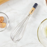 The Zyliss Beech Wood Balloon Whisk, featuring stainless steel wires and a beech wood handle, rests on a marble countertop beside a rolling pin, whole eggs, cracked eggs, and a glass bowl with two cracked eggs.