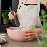 In a modern kitchen with potted herbs, a person in a beige apron mixes ingredients in a large pink ceramic bowl using a heat-resistant Zyliss Spatula with a silicone head. #Medium
