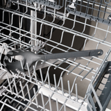 A black spaghetti server utensil is placed inside the bottom rack of an open dishwasher. The dishwasher racks are empty except for the utensil.