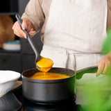 A person in a beige apron stirs orange soup on the stove with a Zyliss Ladle; a white bowl is nearby. #Medium