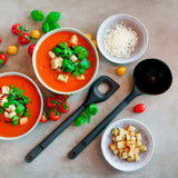 Two bowls of tomato soup topped with croutons and herbs, surrounded by cherry tomatoes, shredded cheese, a bowl of croutons, fresh basil, and two Zyliss Angled Mixing Spoons with ergonomic handles on a light surface. #Large
