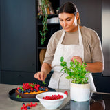 In a modern kitchen, a woman in a beige apron uses the Zyliss Spoontula to decorate a berry-topped cake with mint leaves. Nearby on the counter are red currants in a bowl and a potted mint plant. #Large