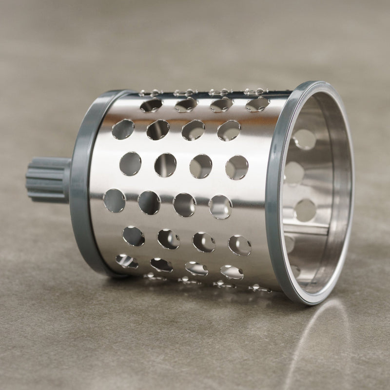 A stainless steel cylindrical grater drum with large round holes and a gray plastic attachment, resting on a smooth, light-colored surface.