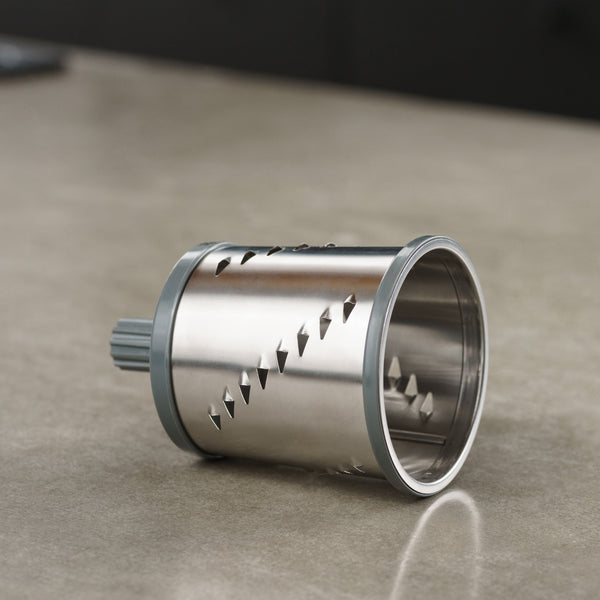 A cylindrical stainless steel rotary grater blade with sharp diagonal slots lies on a smooth, beige countertop. The blade has gray plastic edges and is designed for use in a kitchen appliance.