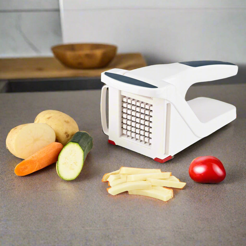 A Zyliss Potato & Vegetable Chipper sits on a kitchen counter with sliced potatoes, carrot, zucchini, tomato, and a wooden bowl in the background. Freshly cut potato strips and its sharp stainless steel blade are visible in front.