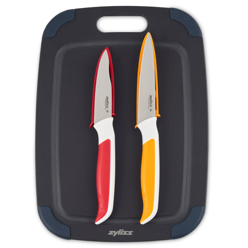 Two kitchen knives with white handles—one red and one orange—are placed side by side on a black cutting board with blue corners and a handle slot at the top.