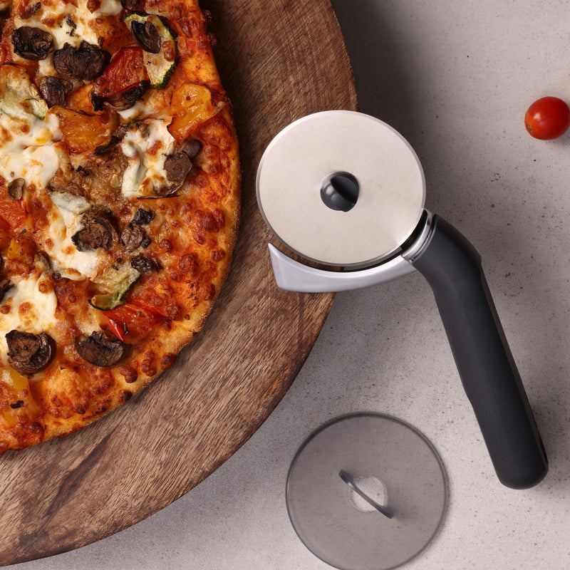 A vegetable pizza on a wooden board sits next to the Zyliss Pizza & Pastry Cutter with ergonomic handle and protective cover on a gray surface. A single cherry tomato is also visible nearby.