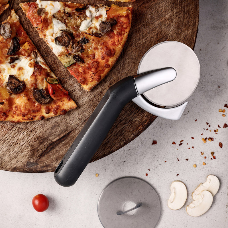 A pizza cutter rests on a white surface next to a sliced pizza on a wooden board. Nearby are pizza toppings including mushroom slices, a cherry tomato, and red pepper flakes.