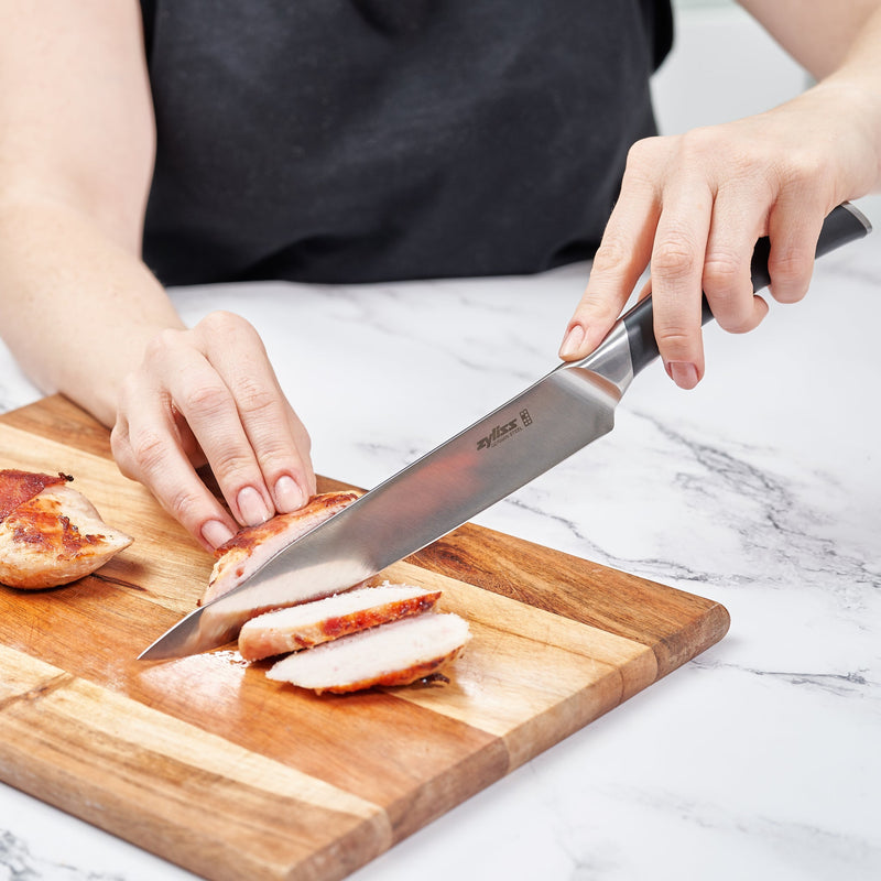 A person slices cooked chicken breast on a wooden cutting board using the Zyliss Comfort Pro Carving Knife 20cm / 8, which features an ergonomic handle, while holding the chicken steady with one hand on a white marble countertop.