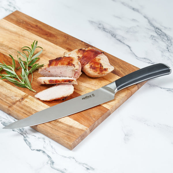 A sliced piece of cooked meat and rosemary are on a wooden board beside the Zyliss Comfort Pro Carving Knife 20cm/8, featuring an ergonomic handle, all arranged on a white marble surface.