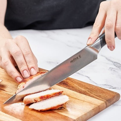 A person slices cooked chicken breast on a wooden cutting board using a large kitchen knife.