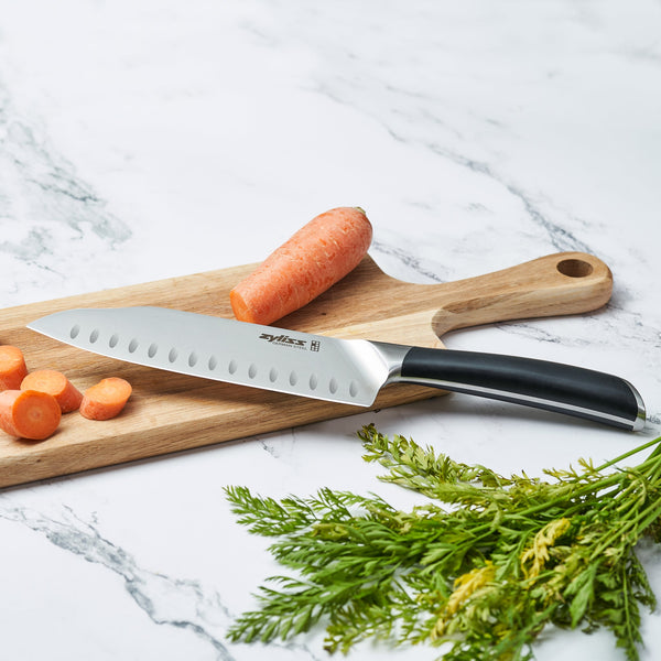 The Zyliss Comfort Pro Santoku Knife 18cm / 7 with an ergonomic handle rests on a wooden cutting board beside carrot slices and leafy tops on a white marble surface.