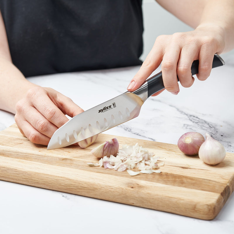 A person chops garlic on a wooden cutting board using the Zyliss Comfort Pro Mini Santoku Knife 13cm / 5 with an ergonomic handle; two whole shallots rest nearby on a white countertop.