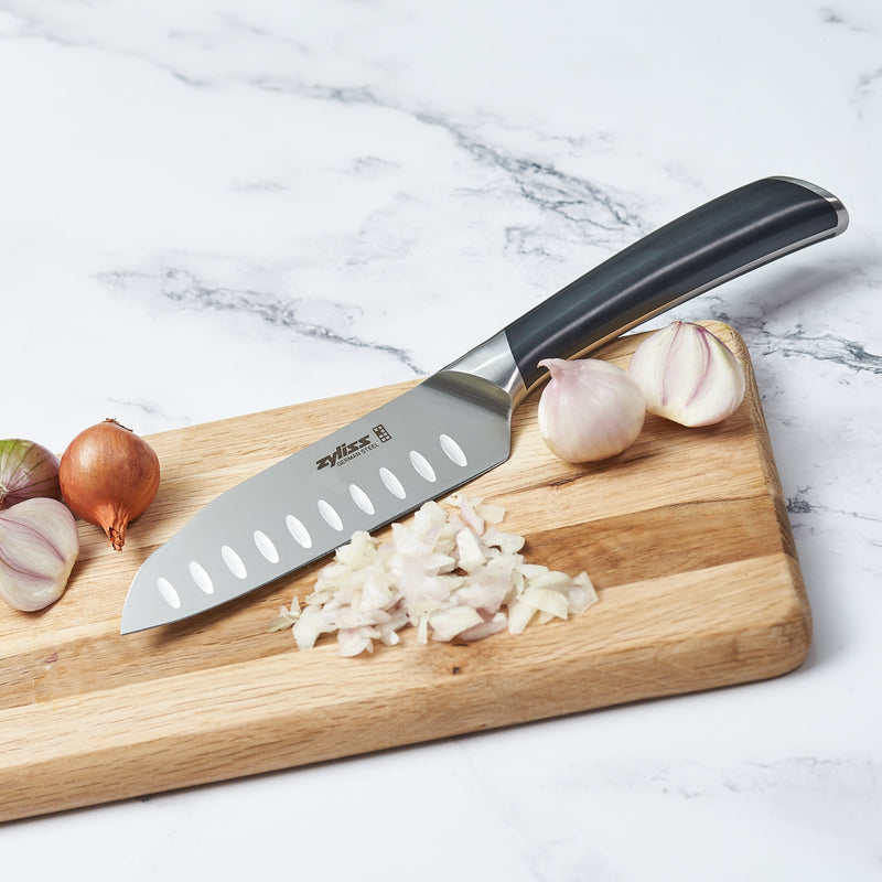 The Zyliss Comfort Pro Mini Santoku Knife 13cm rests on a wooden cutting board next to chopped shallots, whole garlic bulbs, and shallots on a white marble surface.