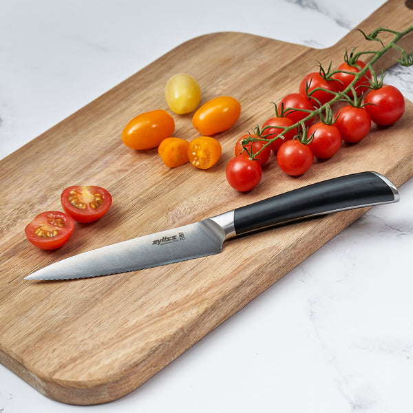 A Zyliss Comfort Pro 3 Piece Paring Knife & Utility Knife Set with black handles is arranged on a wooden cutting board with assorted whole and sliced red and yellow cherry tomatoes, all atop a white marble surface.