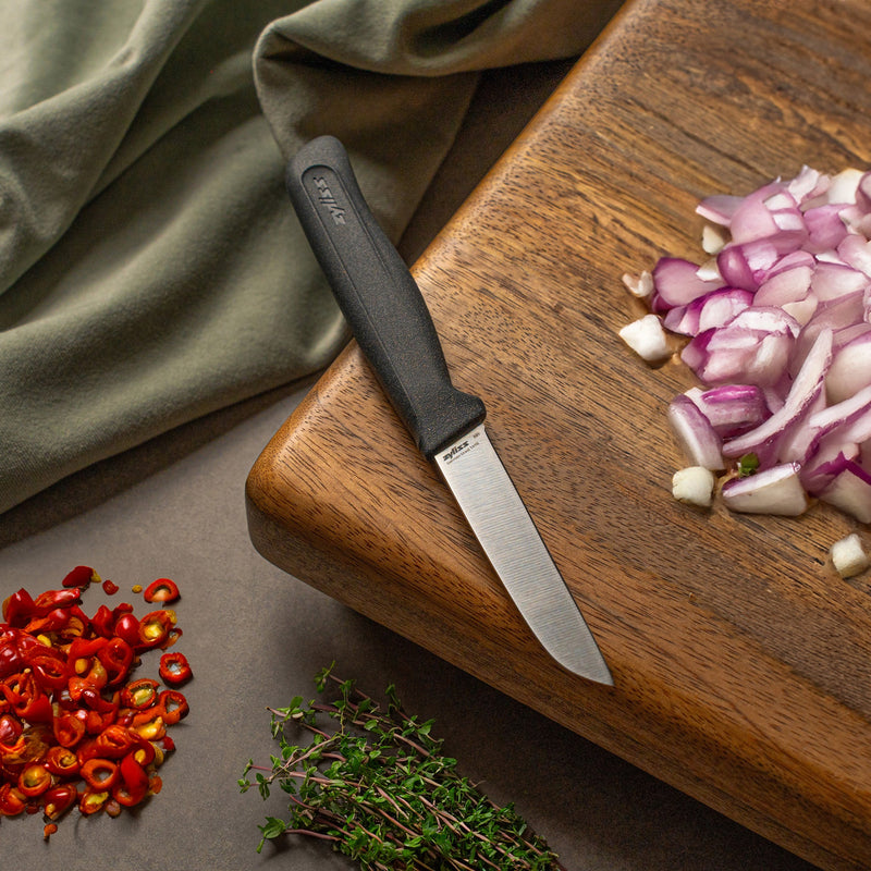 A Zyliss Vegetable Knife 9cm sits on a wooden cutting board next to chopped red onions, with sliced red chili peppers and fresh thyme close by. A green cloth in the background adds a pop of color.