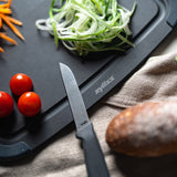 A Zyliss knife rests on a black cutting board beside sliced cucumber, carrot strips, cherry tomatoes, and a loaf of bread on a beige cloth.