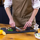 A person in a brown apron slices a cooked steak on a black cutting board, with chopped herbs, mushrooms, a lemon, and a pepper grinder nearby on a wooden table.