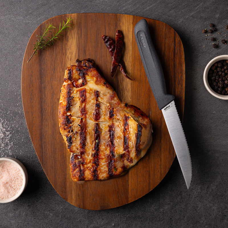 A grilled steak with dark grill marks rests on a wooden board next to a Zyliss Steak Knife Serrated 11cm, rosemary, two dried red chilies, and bowls of black peppercorns and pink salt.