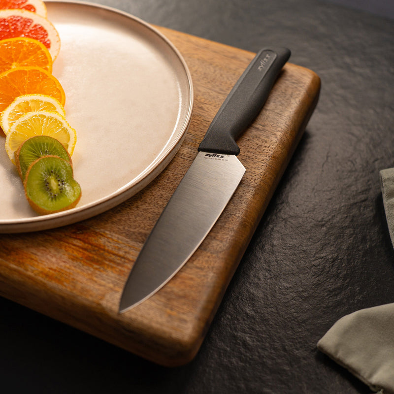 #15cm/6"
The Zyliss Chef Knife 15cm / 6 with an ergonomic handle lies on a wooden cutting board beside a plate of sliced citrus and kiwi, all arranged on a dark countertop.