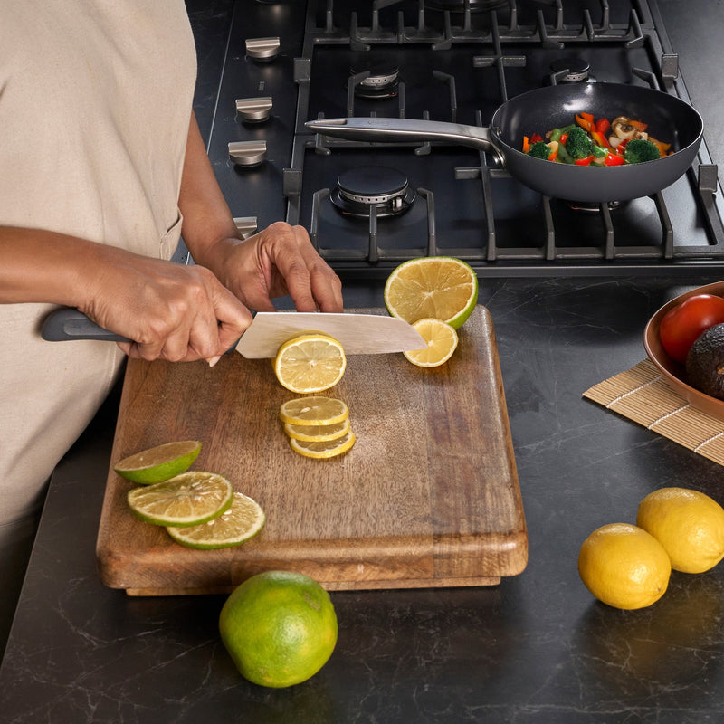 #17cm/7"
A person uses a Zyliss Santoku Knife with an ergonomic handle to slice a lemon on a wooden cutting board next to limes and whole lemons, while vegetables cook in a pan on the stove in the background.