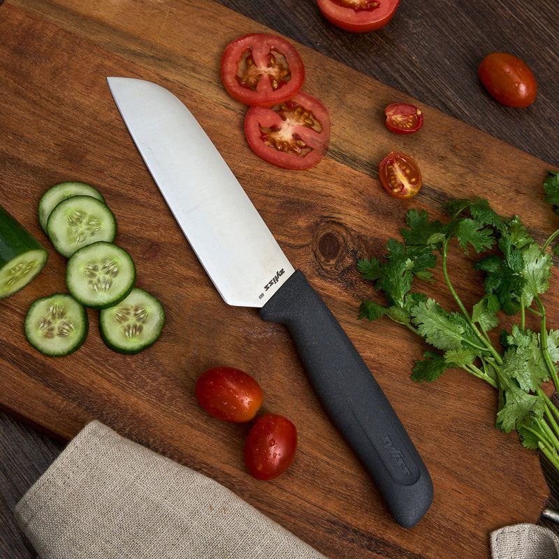 #17cm/7"
A Zyliss Santoku Knife with an ergonomic handle rests on a wooden cutting board, surrounded by sliced cucumbers, tomatoes, cilantro, and a beige towel atop a dark table.