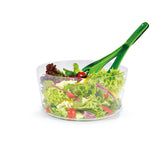 A clear bowl filled with mixed green salad, cherry tomatoes, and red onion slices, with a pair of green salad servers resting inside.