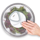 A hand presses a button on the lid of the Zyliss Easy Spin 2 Stainless Steel Salad Spinner, which has a non-slip base and is filled with leafy greens, seen from above.