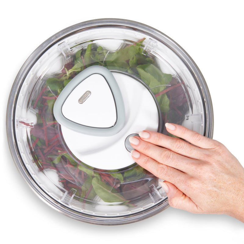 A hand presses a button on the lid of the Zyliss Easy Spin 2 Stainless Steel Salad Spinner, which has a non-slip base and is filled with leafy greens, seen from above.