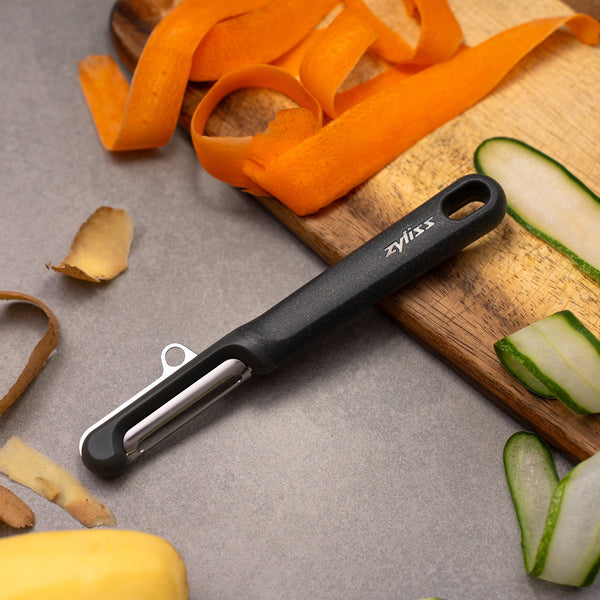 A Zyliss Swivel Peeler with a black ergonomic handle rests on a gray surface near a wooden cutting board, surrounded by strips of peeled carrot, cucumber, and potato skins.