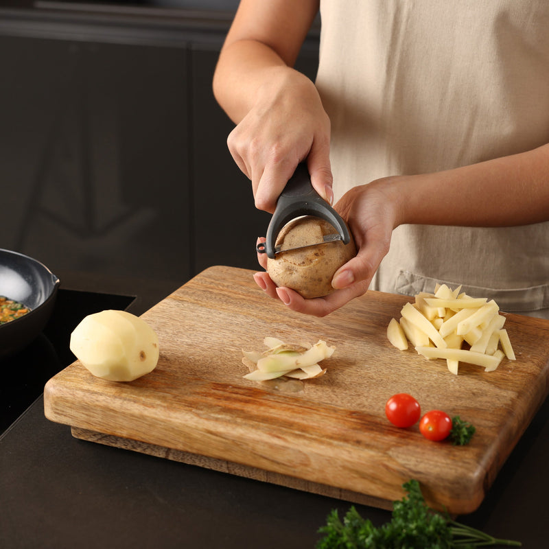 A person peels a potato with the Zyliss Y Peeler, which has a stainless steel swivel blade and ergonomic handle, on a wooden cutting board with peeled potato, slices, cherry tomatoes, and parsley around.
