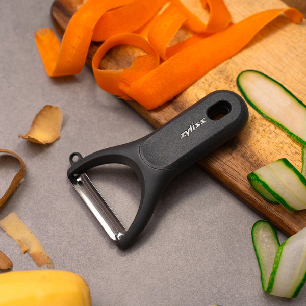 A Zyliss Y Peeler with a black ergonomic handle rests on a gray surface scattered with peeled carrot, cucumber, and potato skins, while a wooden cutting board displays additional colorful vegetable peels.