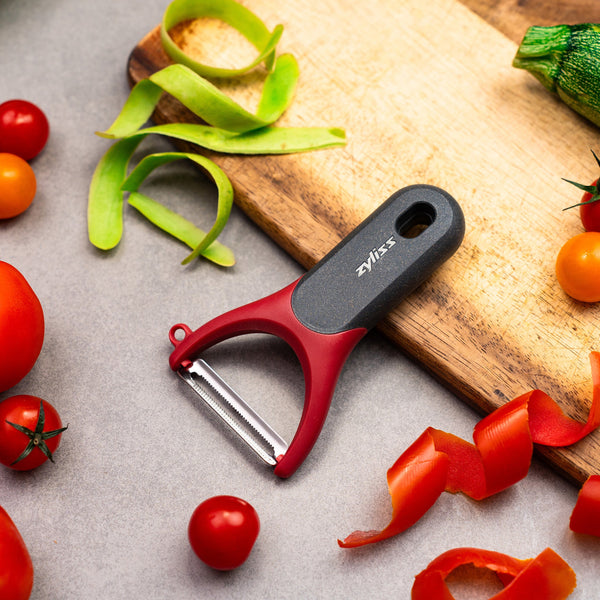 A red and black Zyliss Soft Skin Y Peeler with an ergonomic grip rests on a gray surface, surrounded by cherry tomatoes, tomato skins, green peels, and a wooden cutting board holding a partially sliced zucchini.