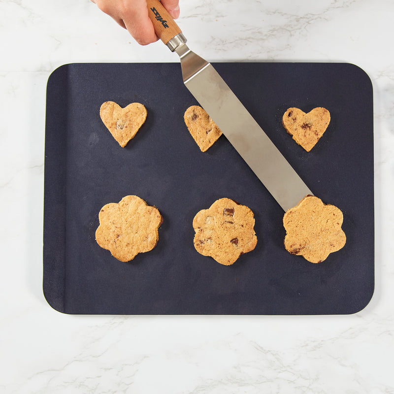 A hand uses the Zyliss Beech Wood Angled Palette Knife to lift a flower-shaped chocolate chip cookie from a baking sheet holding three flower and three heart cookies on a white countertop.