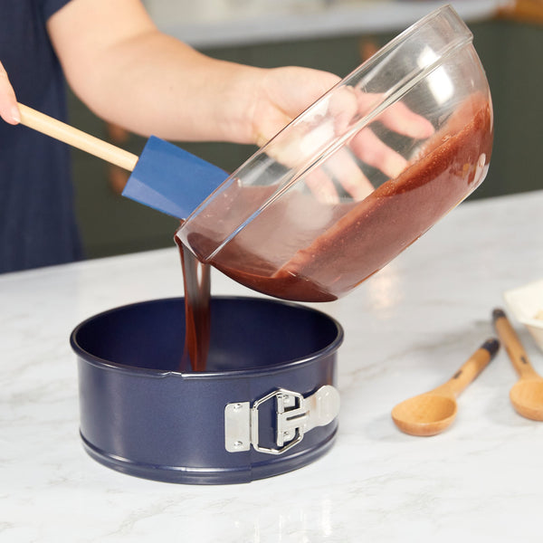 Someone pours cake batter from a glass bowl into the Zyliss Durable Non-Stick Springform Cake Pan using a blue spatula on a light countertop, with wooden spoons nearby. #20cm/8"