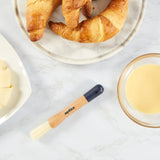 On a marble surface: croissants on a plate, a bowl of egg wash, and the Zyliss Beech Wood Pastry Brush with natural bristles beside unbaked dough on a white plate.