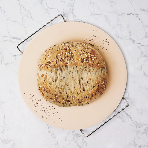 A round loaf of multigrain bread with seeds on top sits on a light-colored plate atop a wire rack, set against a white marble background.