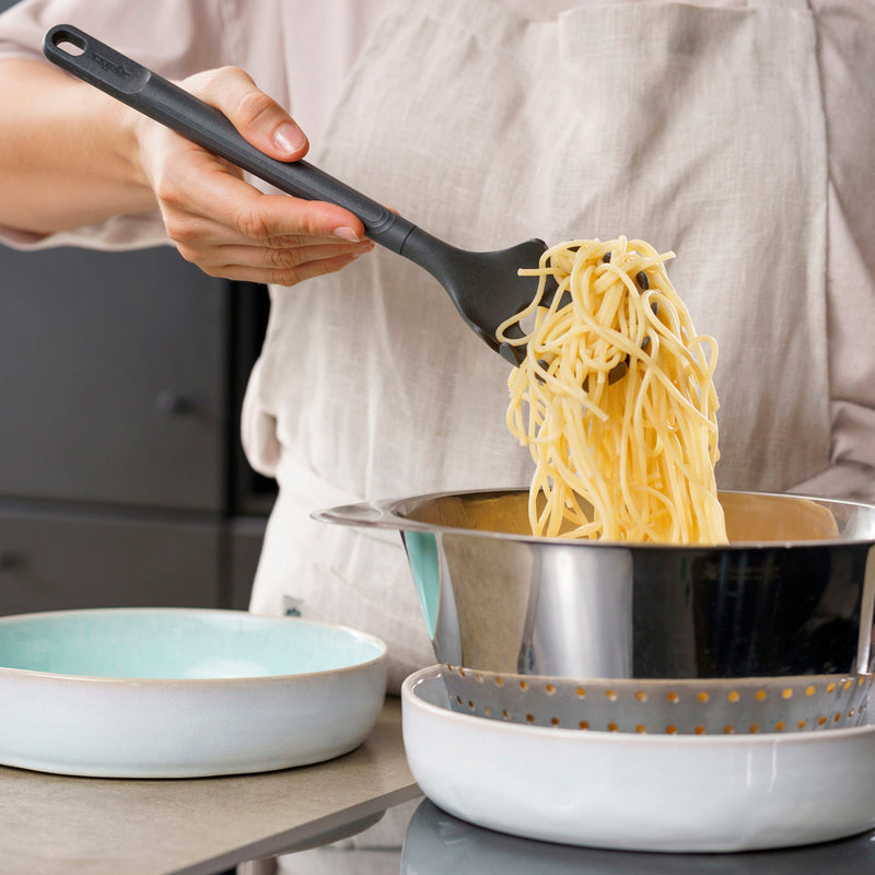 A person in a light apron uses a Zyliss Spaghetti Server to lift cooked spaghetti from a metal colander over a pot, with a light blue bowl on the kitchen counter nearby.