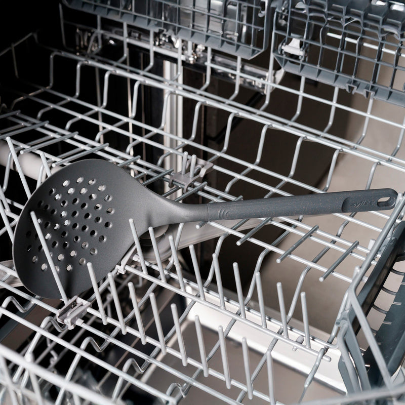 A large black slotted spoon rests diagonally across the top rack of an open dishwasher, surrounded by metal prongs designed to hold dishes.