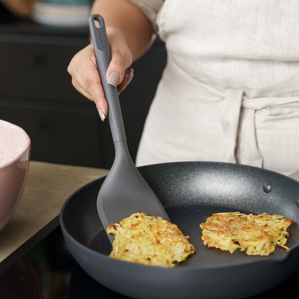 A person in a beige apron uses a spatula to cook two potato pancakes in a black nonstick frying pan on a stovetop.