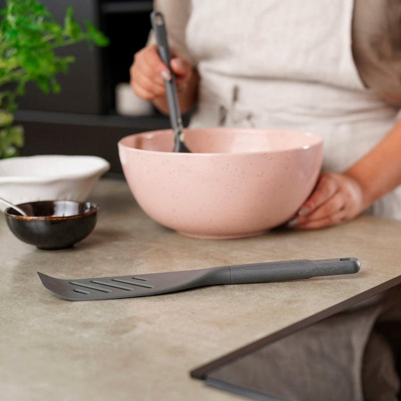 A person in an apron stands at the kitchen counter, mixing ingredients in a large pink bowl. A Zyliss Fish Turner rests nearby, along with a small white bowl and a plant visible in the background.