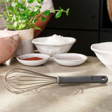 A Zyliss Balloon Whisk with a gray silicone handle sits on a kitchen counter, safe for non-stick cookware. It’s surrounded by bowls of flour and cocoa powder, with a plant and part of a person’s arm in the background. #Large