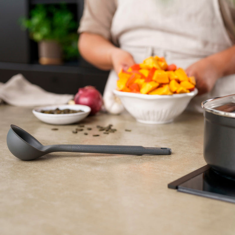 A person in an apron holds a bowl of chopped orange squash on a kitchen counter, with a Zyliss Ladle, a pot, a red onion, and a small bowl of seeds nearby. #Medium