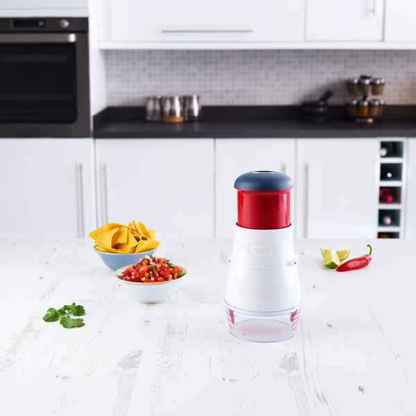 The Zyliss Zick-Zick 2 Food Chopper with a blade cover sits on a kitchen counter next to tortilla chips, salsa, a lime wedge, red chili pepper, and cilantro in a modern kitchen.